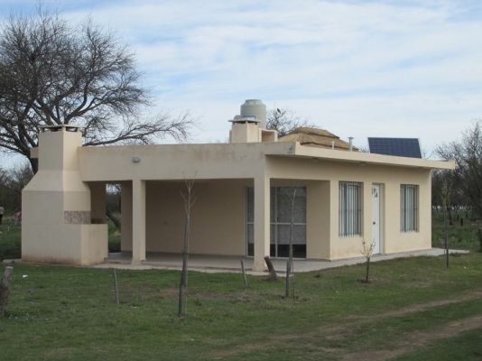 Casa de una sola planta color beige con patio y barbacoa en un campo de hierba bajo un cielo nublado.