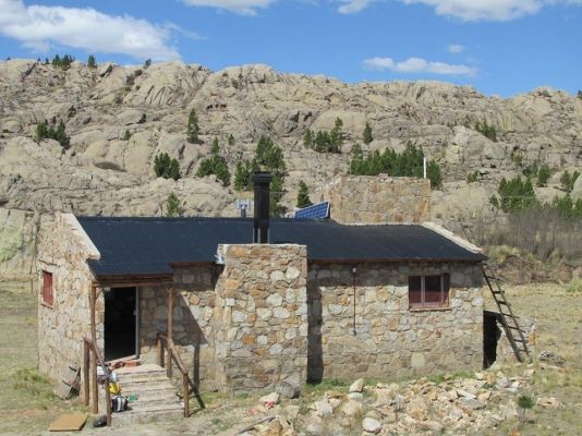 Cabaña de piedra con techo oscuro y chimenea, situada contra una ladera rocosa y un cielo azul.