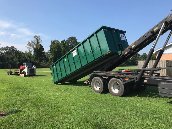 A green dumpster is being loaded onto a trailer in a grassy field.