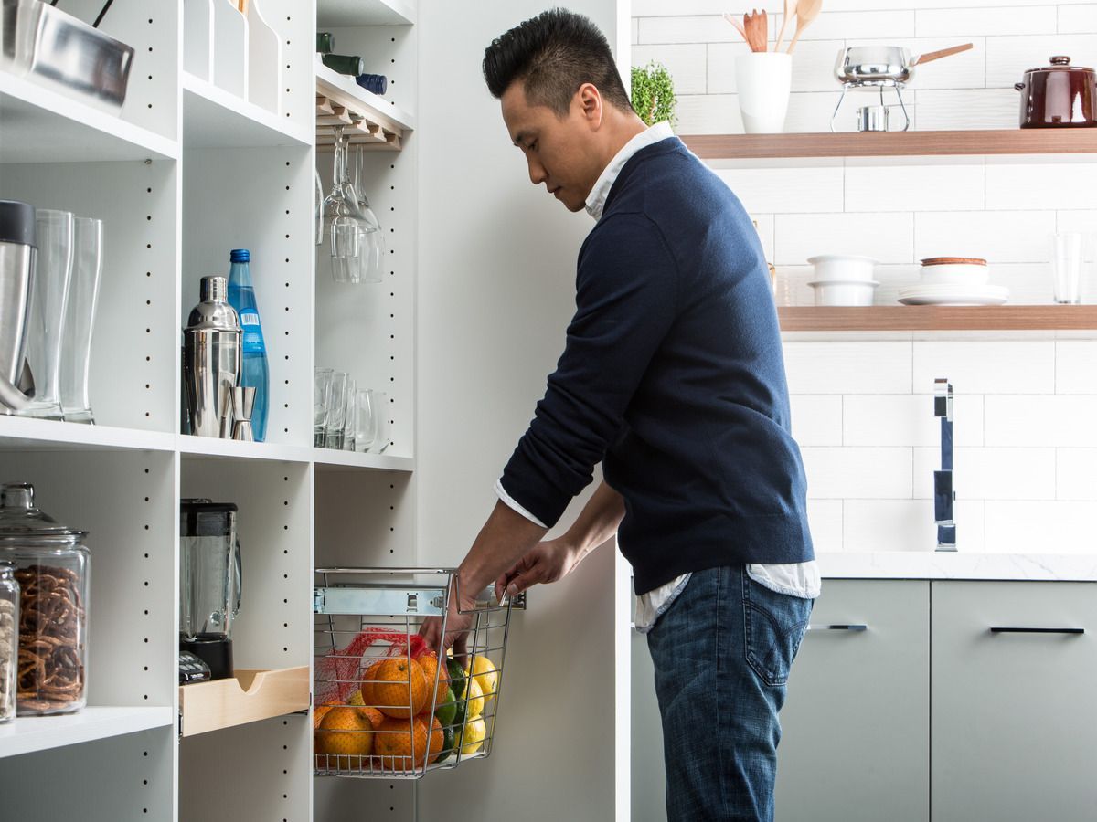 A man is pulling a basket of fruit out of a cabinet in a kitchen.