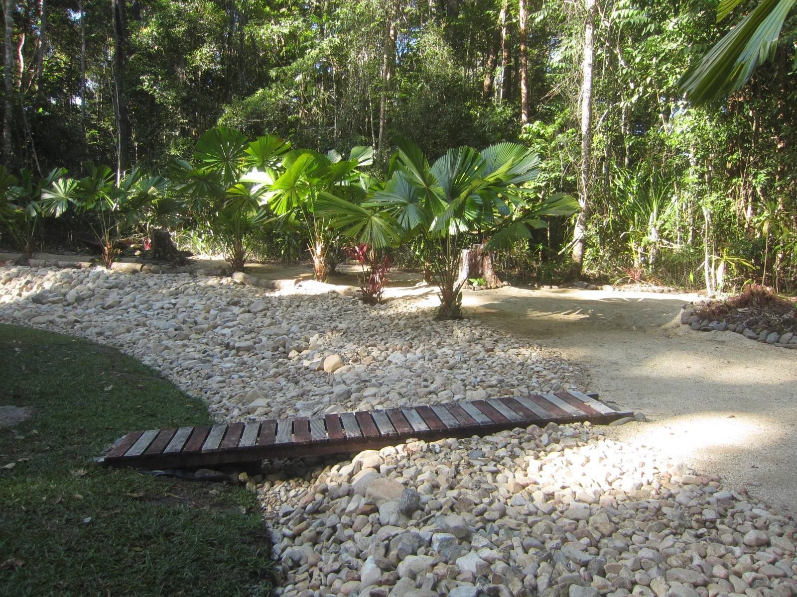 Wooden bridge over gravel pathway in a lush garden with trees — Kuranda Landscape Supplies in Kuranda, QLD