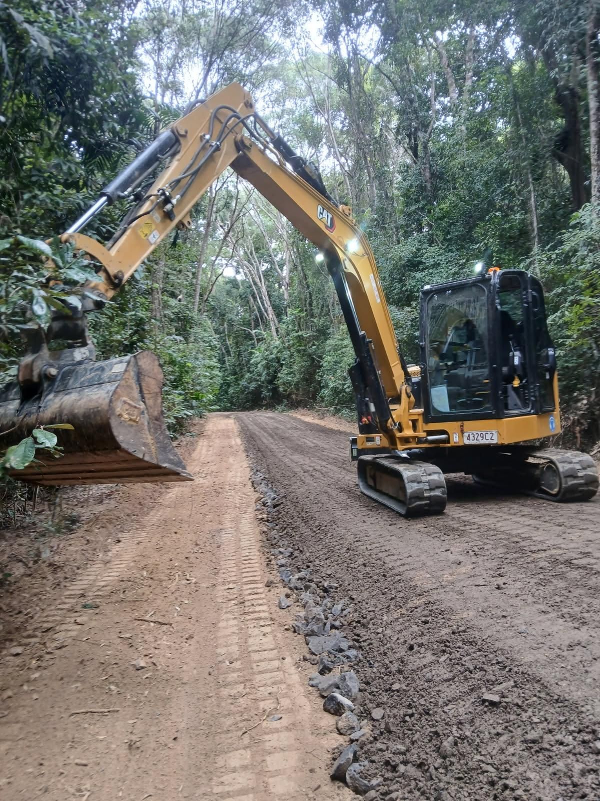 Yellow excavator clearing a dirt road in a forest, removing a fallen log — Kuranda Landscape Supplies in Kuranda, QLD