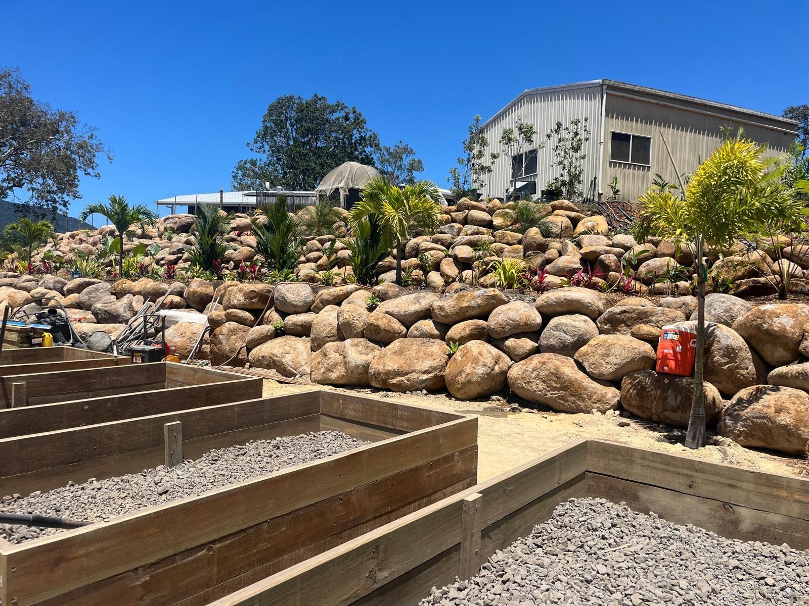 Raised garden beds in foreground, large rock wall and building in the background on a sunny day — Kuranda Landscape Supplies in Kuranda, QLD