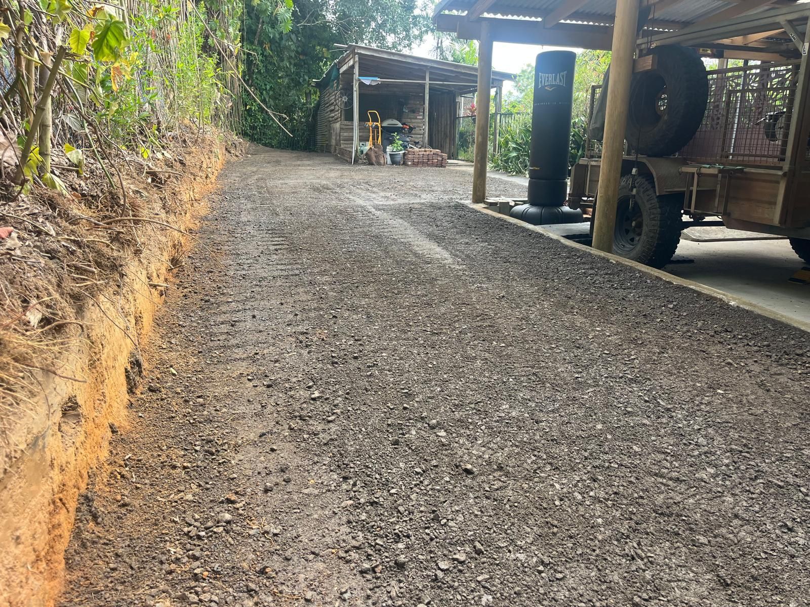A gravel driveway next to a dirt embankment, leading toward a shed. A trailer sits on the right — Kuranda Landscape Supplies in Kuranda, QLD