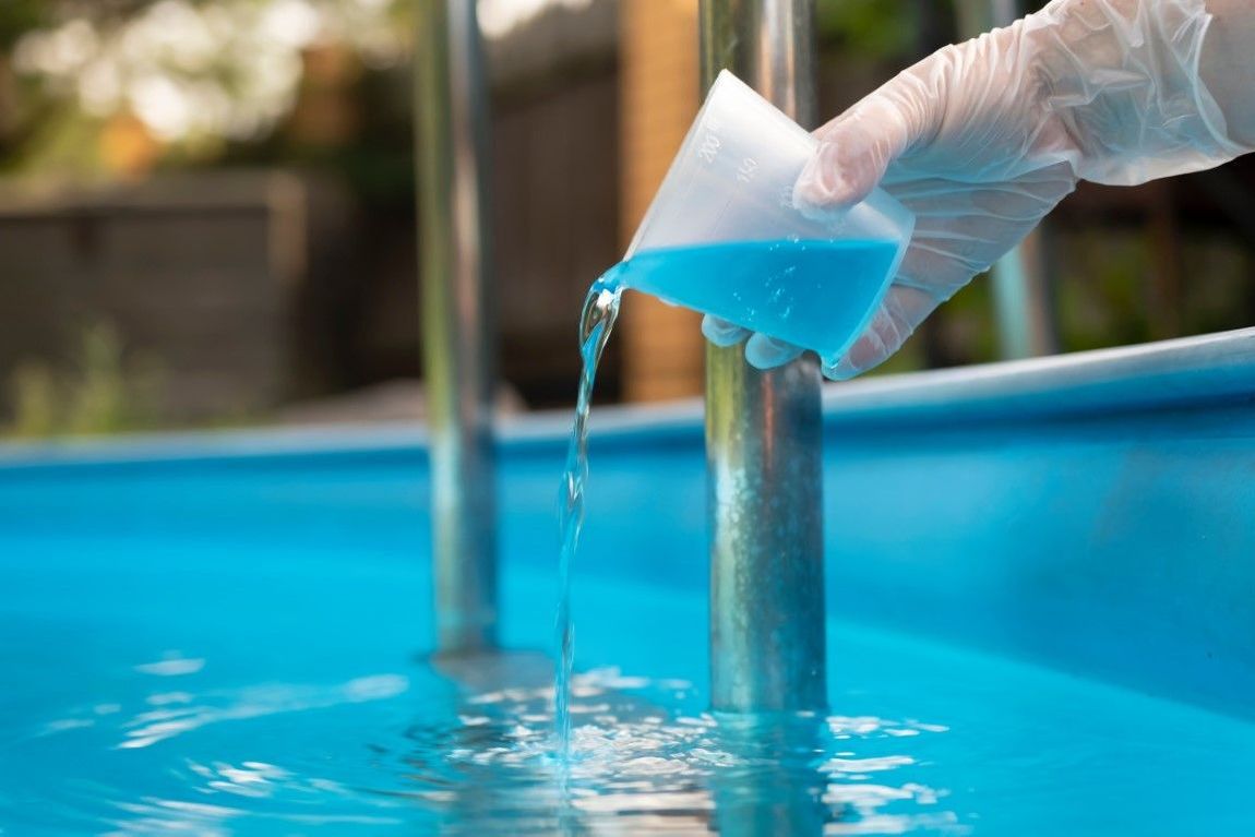A gloved hand pouring blue liquid from a cup into a swimming pool.