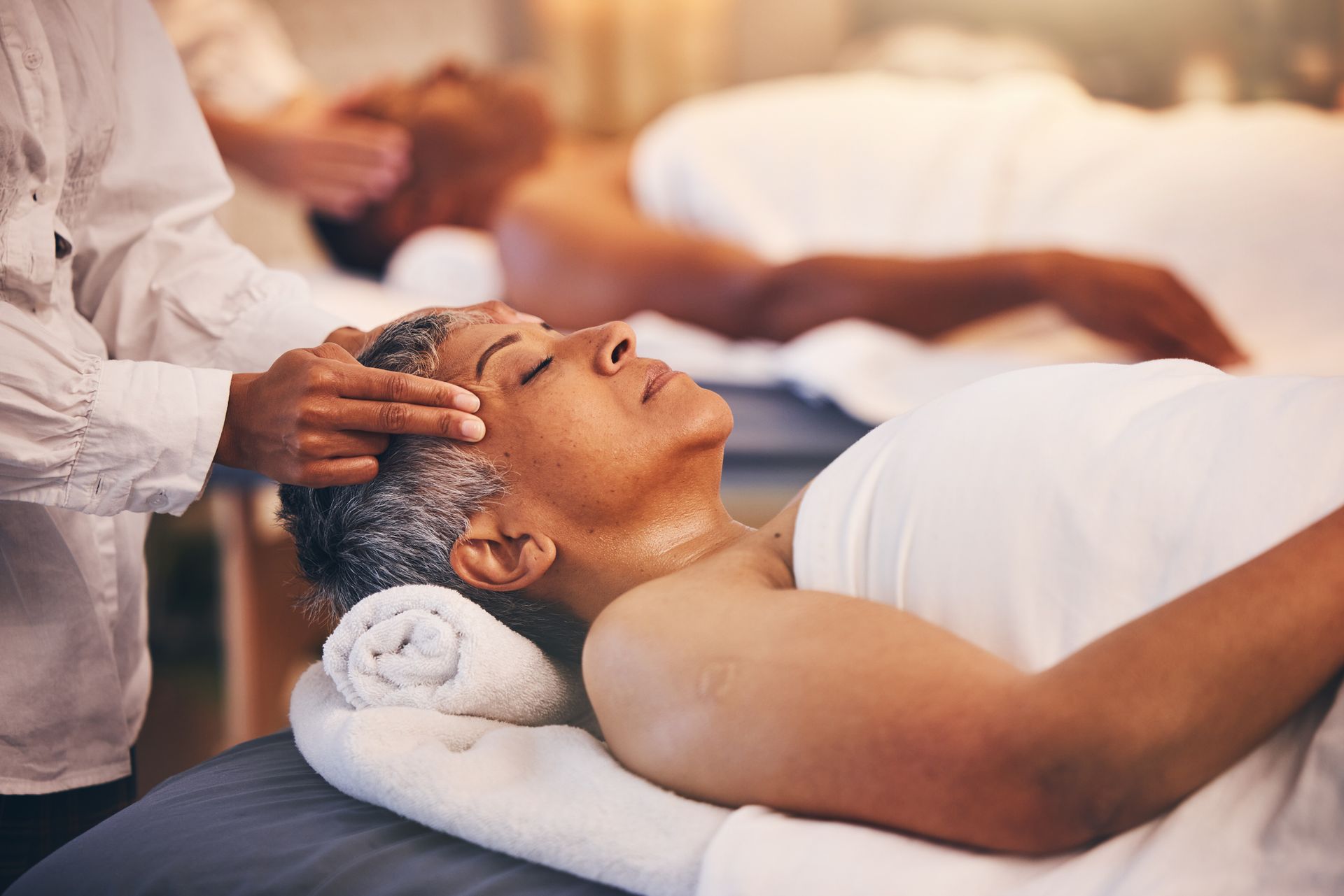 Person receiving a head massage; lying on a massage table with eyes closed, other person in background.