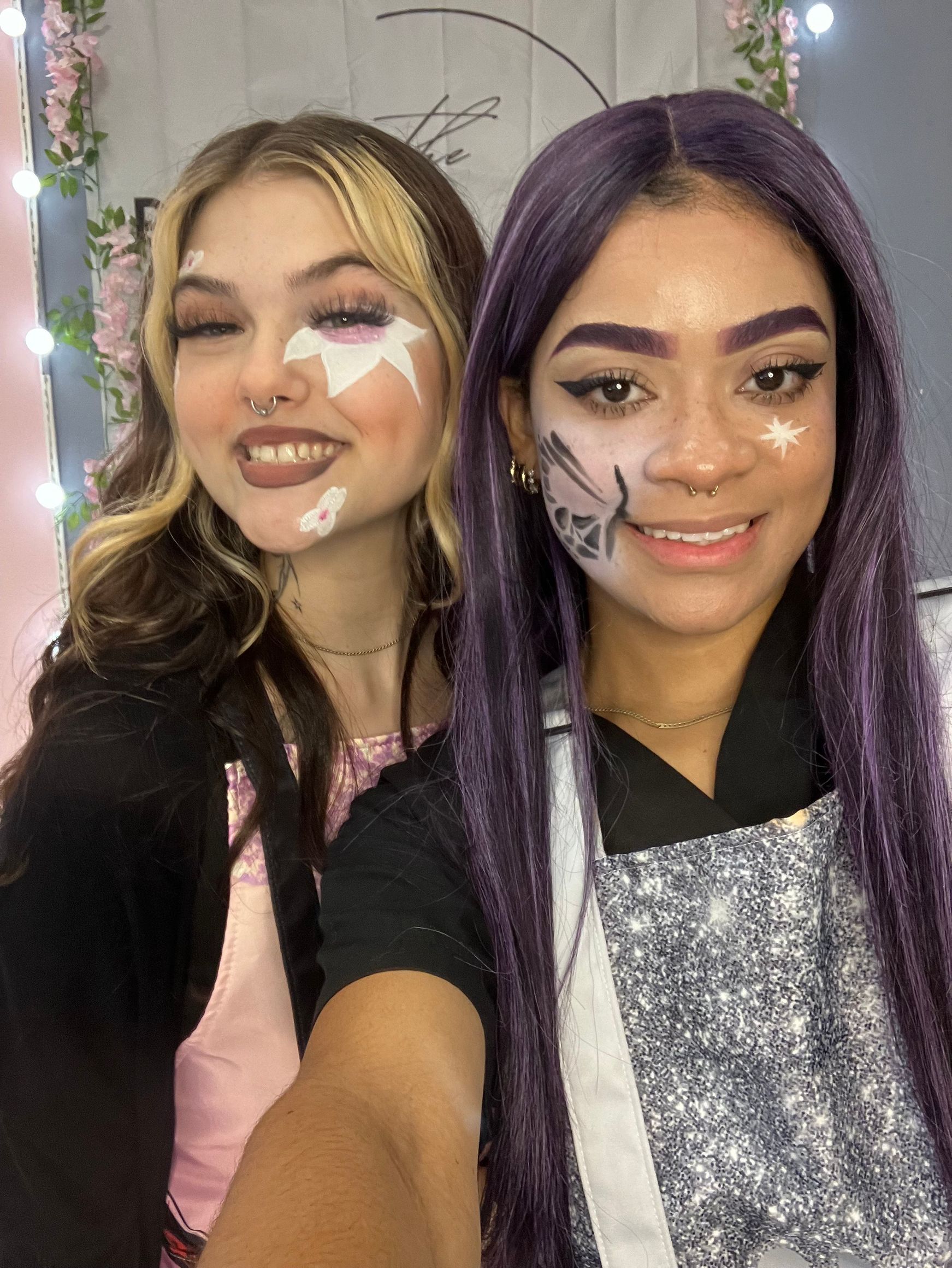 Two smiling people with face makeup, one with blonde hair, the other with purple. Both are indoors.