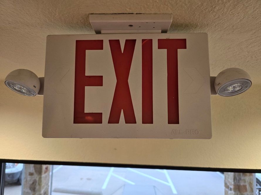 Green fire exit sign, white lettering, man running toward a downward arrow, mounted on a white wall.