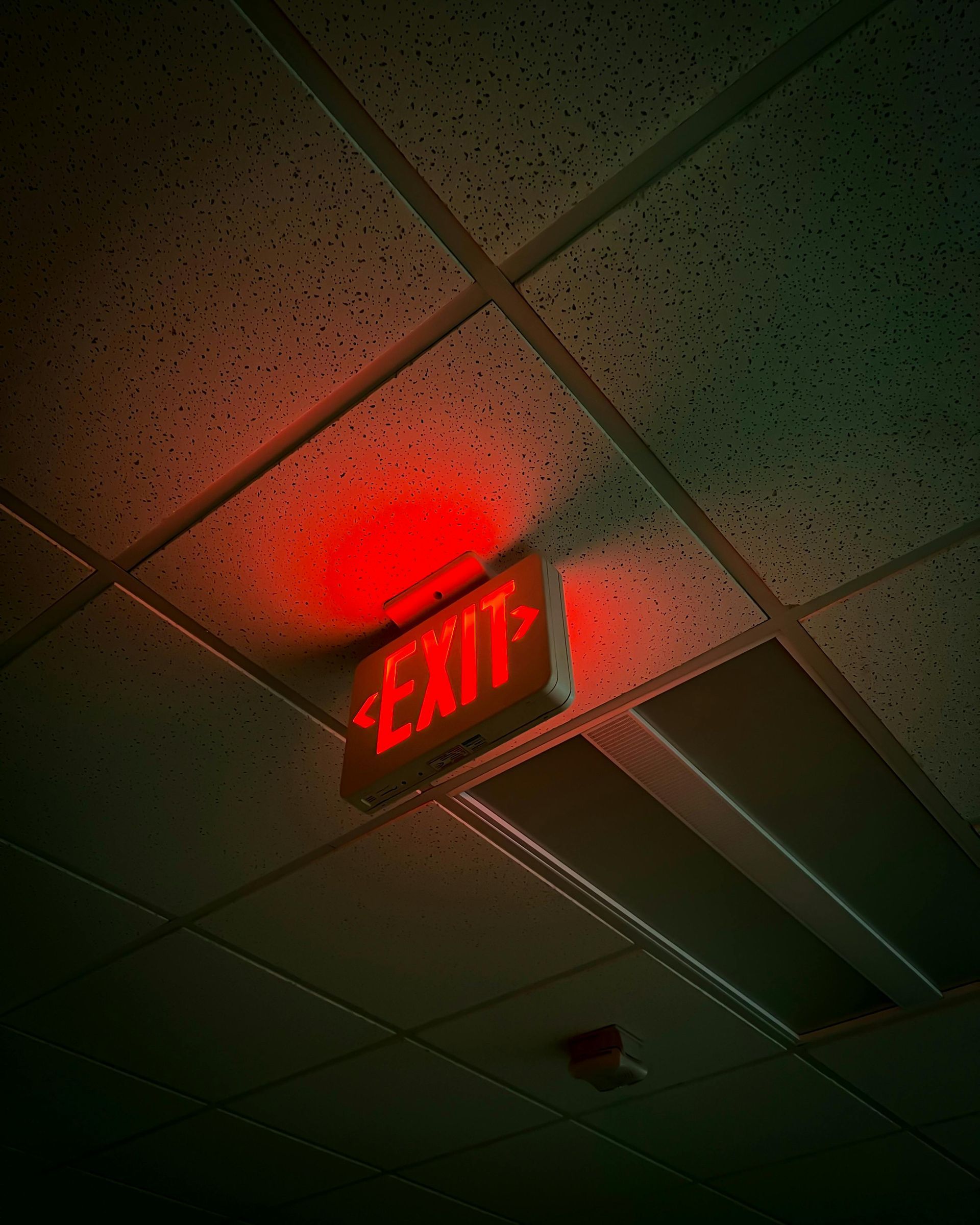 A glowing red EXIT sign hangs from a dark, tiled ceiling, illuminating the surrounding area with a faint red light.