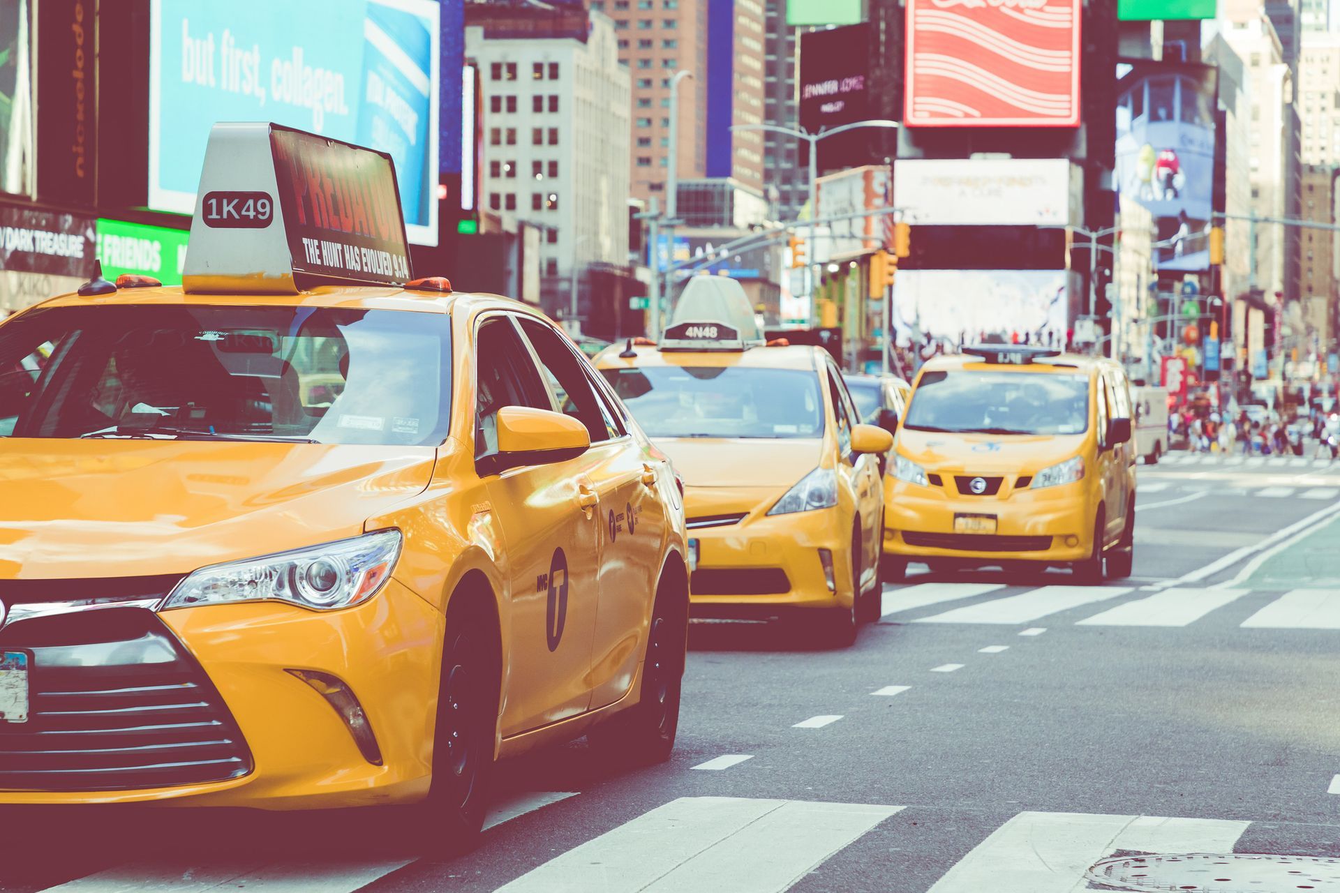 Yellow New York City taxis on a busy street with crosswalks, buildings, and billboards.