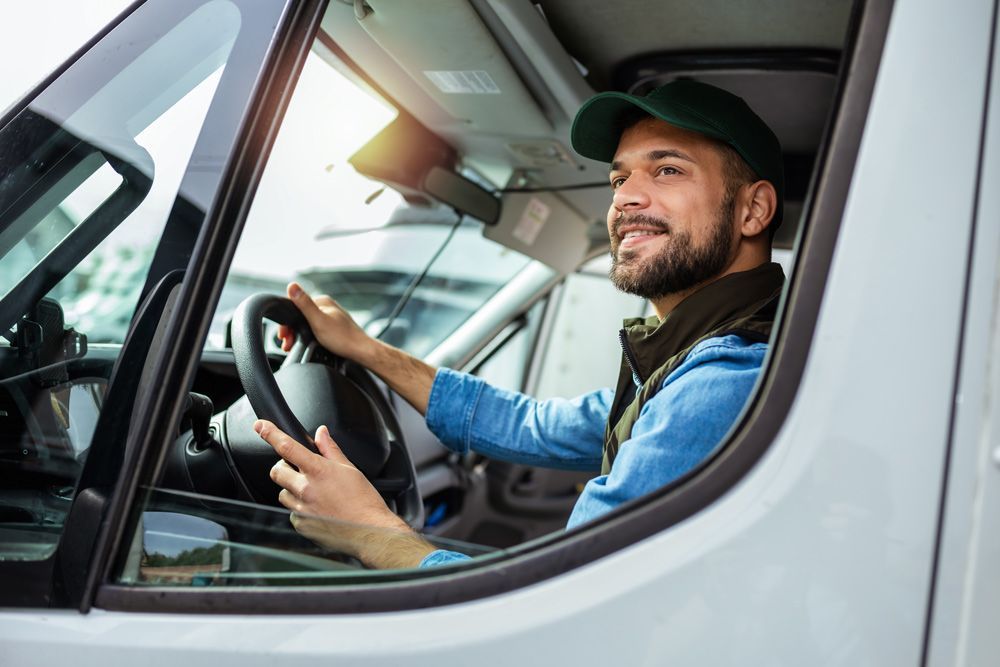 Man driving a white vehicle, smiling, wearing a green cap and blue shirt.