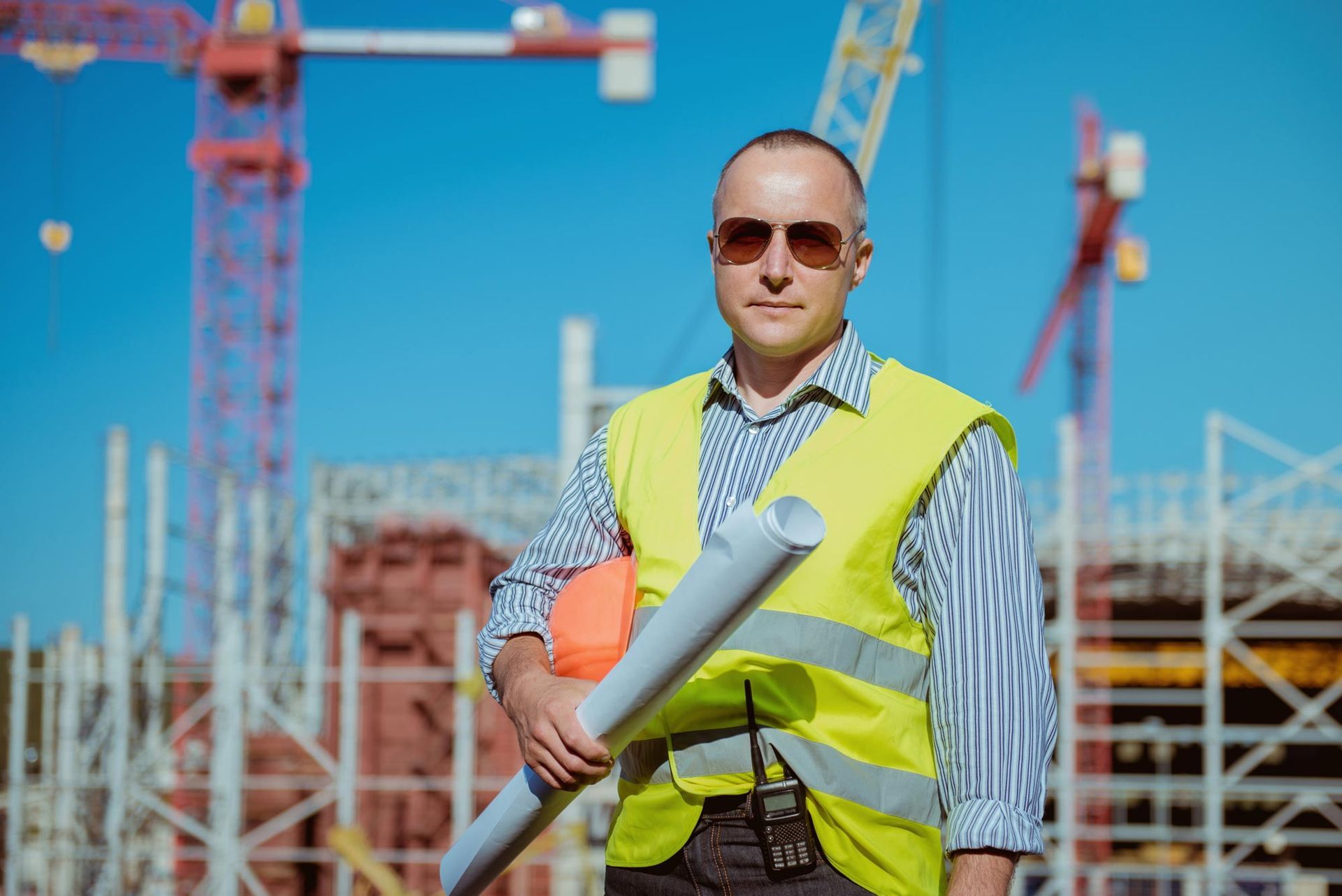 Construction worker holding plans and helmet, standing in front of a building site with cranes.