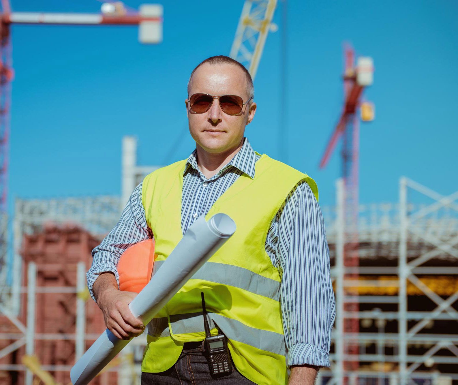 Construction worker holding blueprints, wearing a vest and sunglasses, standing in front of construction site with cranes.