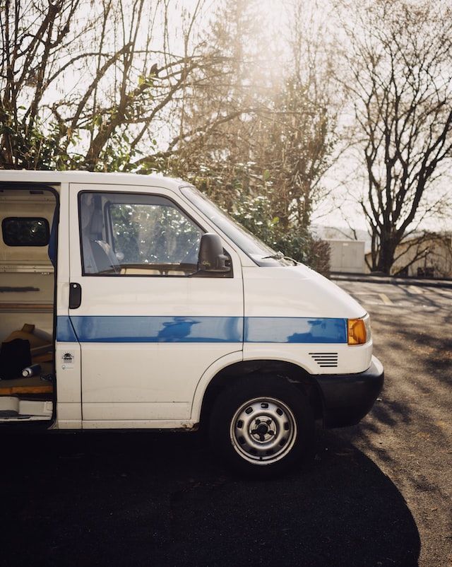 White van with blue stripe parked outdoors, door open. Sunlight shines behind trees.