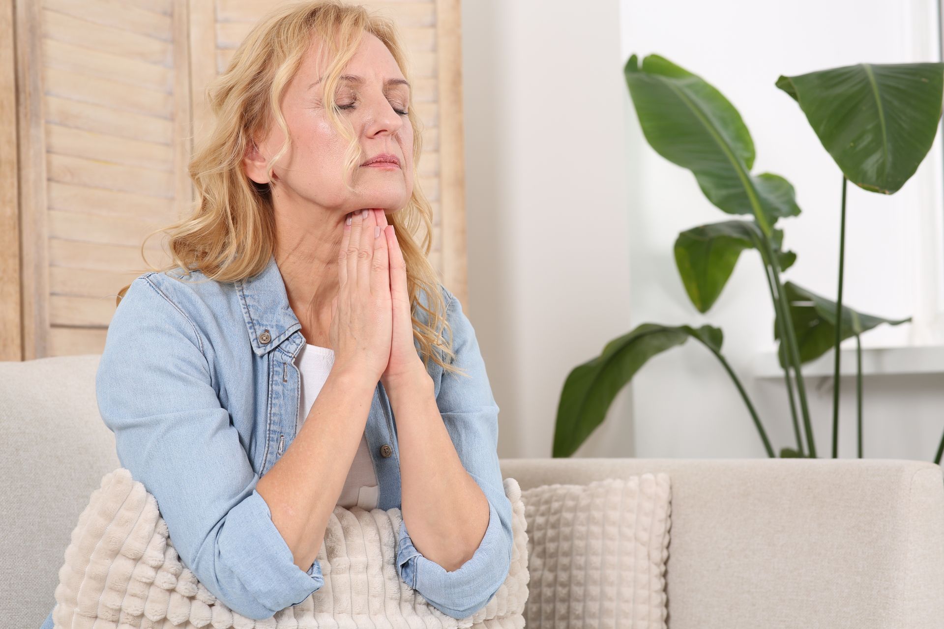 A tired middle-aged woman sits on a couch with her eyes closed and hands pressed together, reflectin