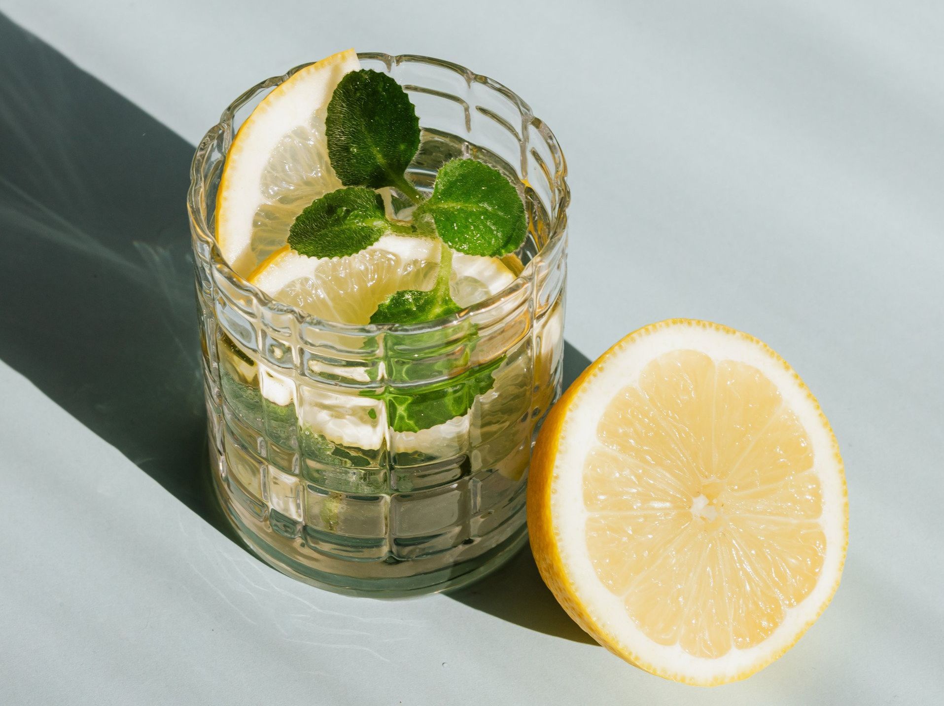 A glass of water with lemon slices and mint leaves next to a slice of lemon.