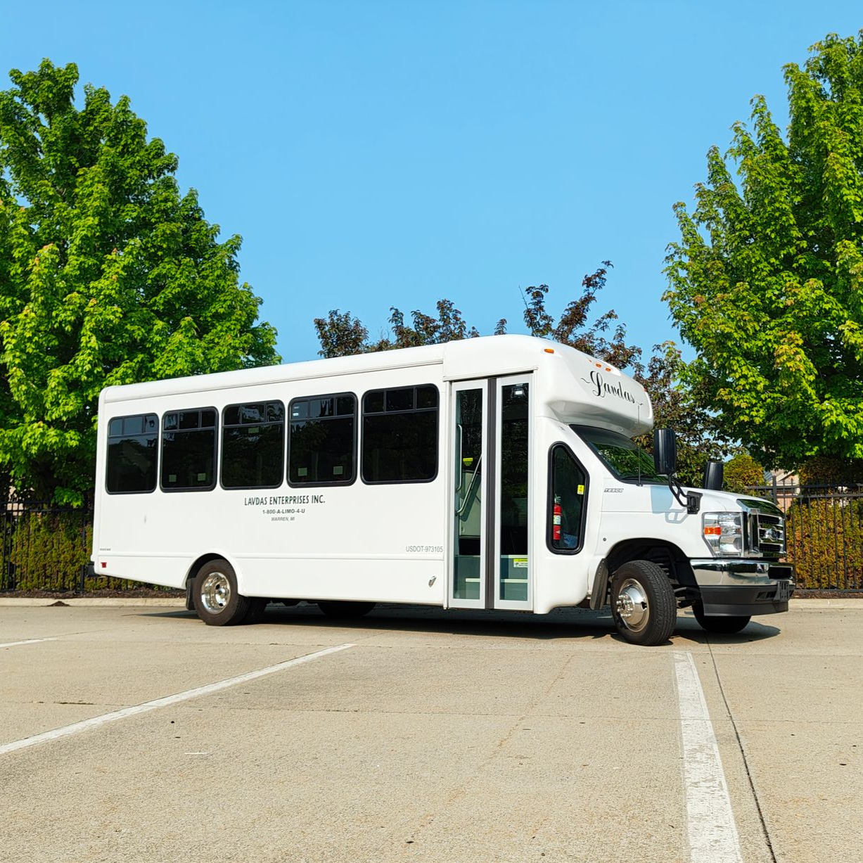 White shuttle bus parked on asphalt, green trees in background, clear blue sky.