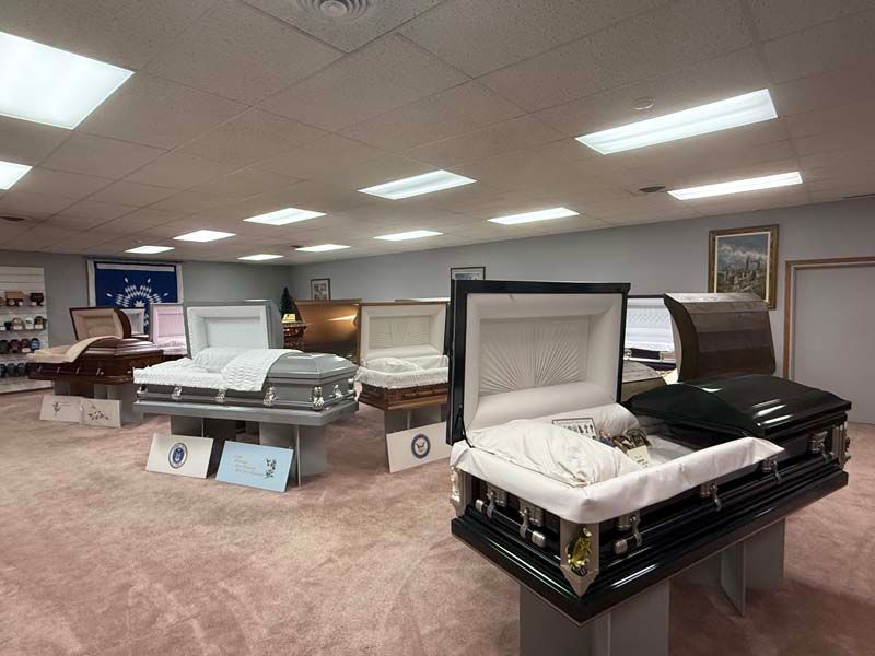A room displaying multiple open and closed caskets on stands; light-colored carpet and walls, fluorescent lights.