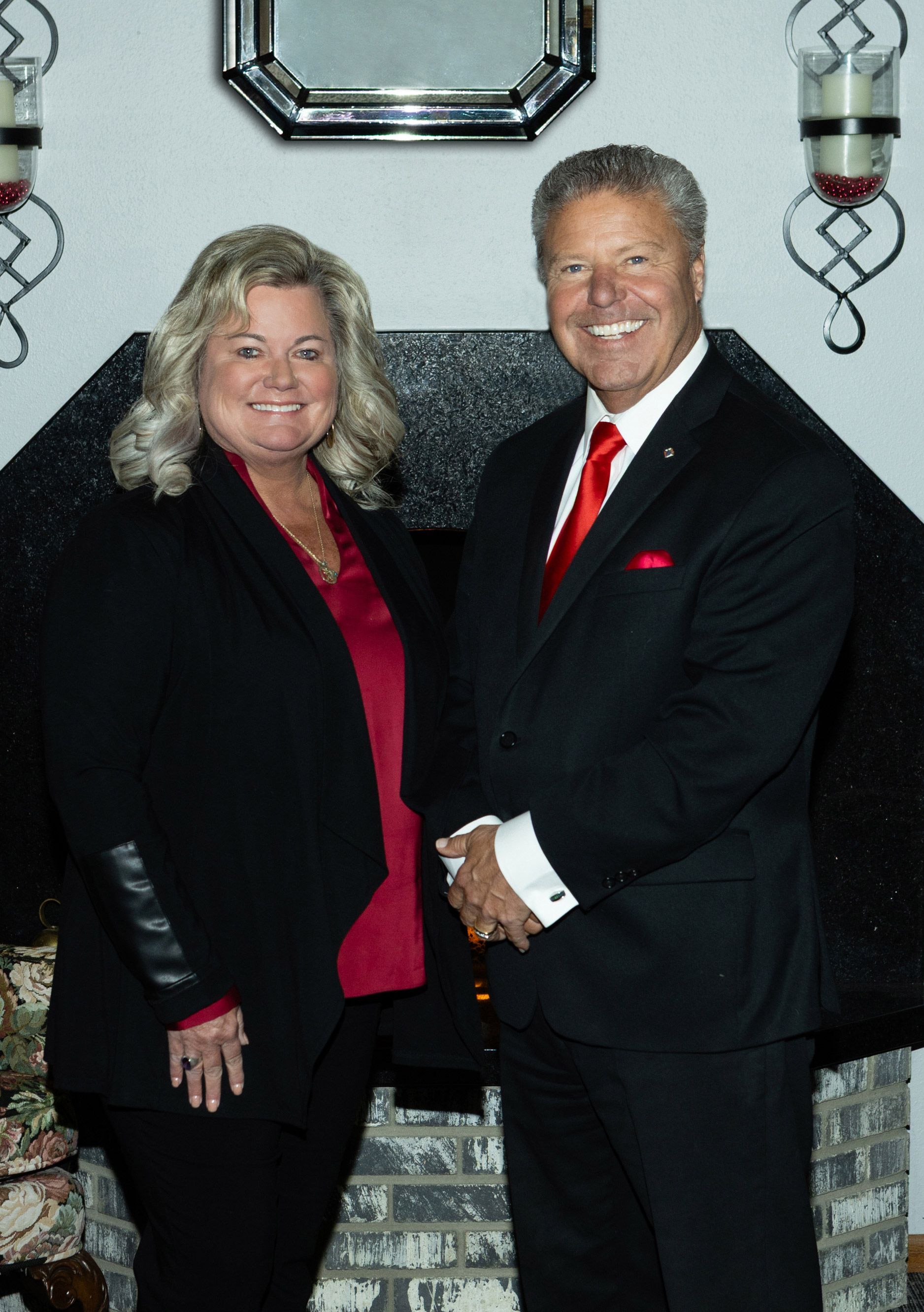 Woman and man standing near a fireplace, smiling. Woman in black jacket, red top. Man in black suit, red tie.