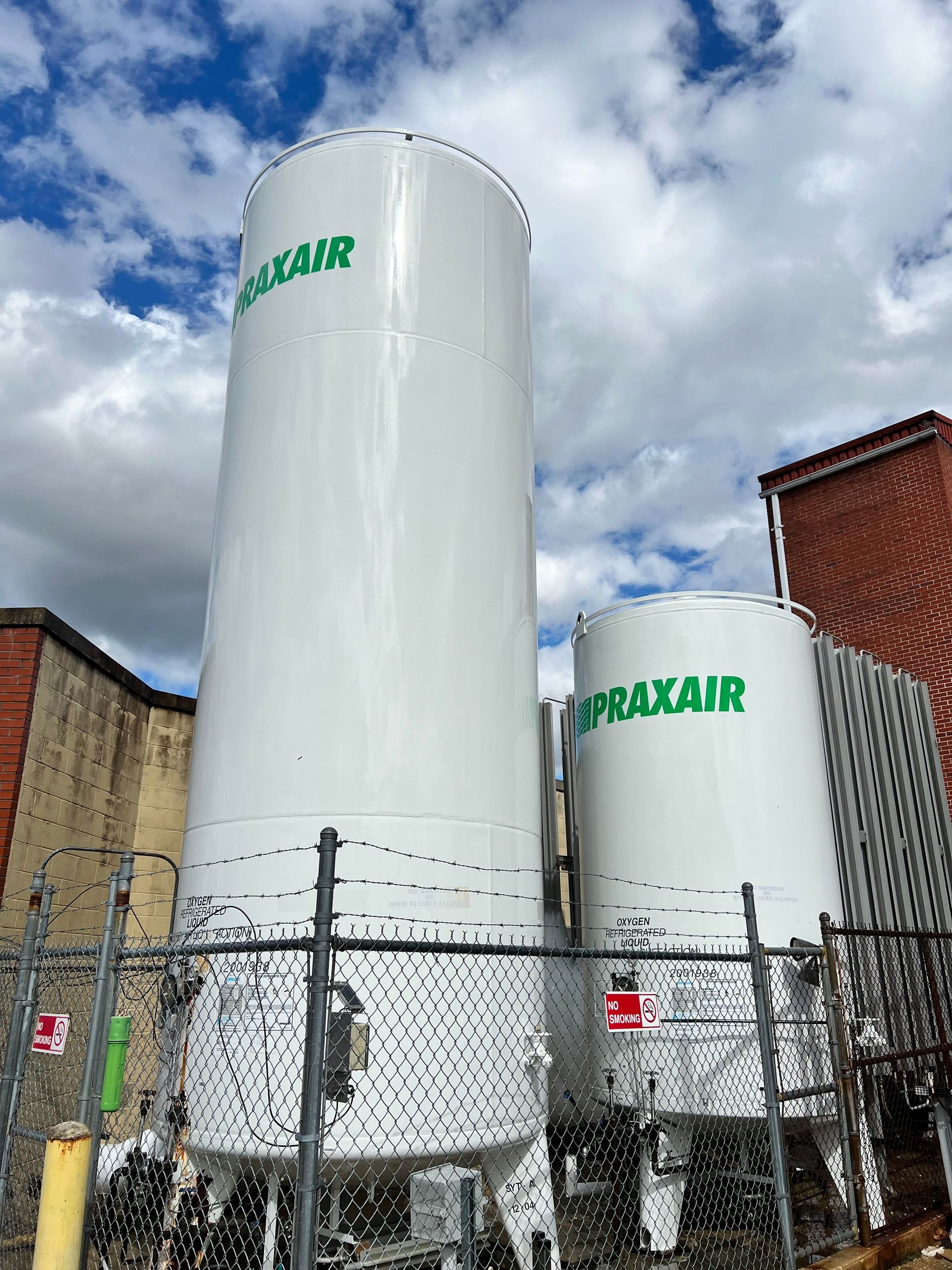 Two large white tanks with the word praxair on them are behind a chain link fence.