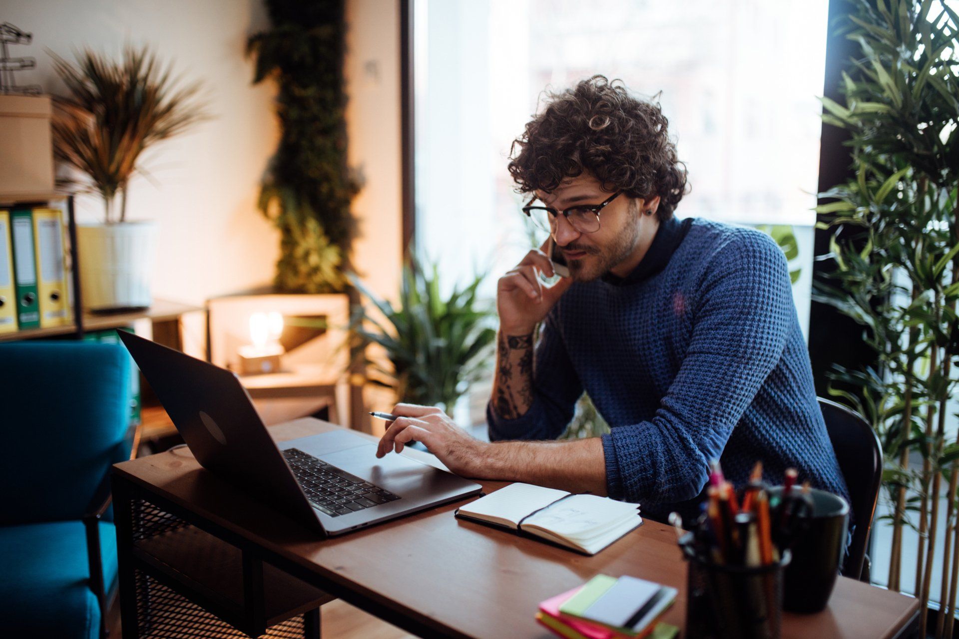 A man is sitting at a desk using a laptop and talking on a cell phone.