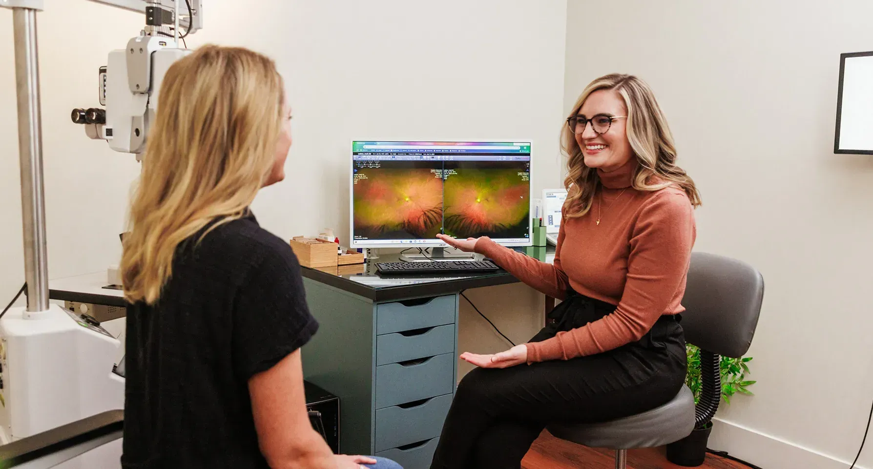 Optometrist pointing at a retinal image on a monitor, explaining it to a patient in an exam room.