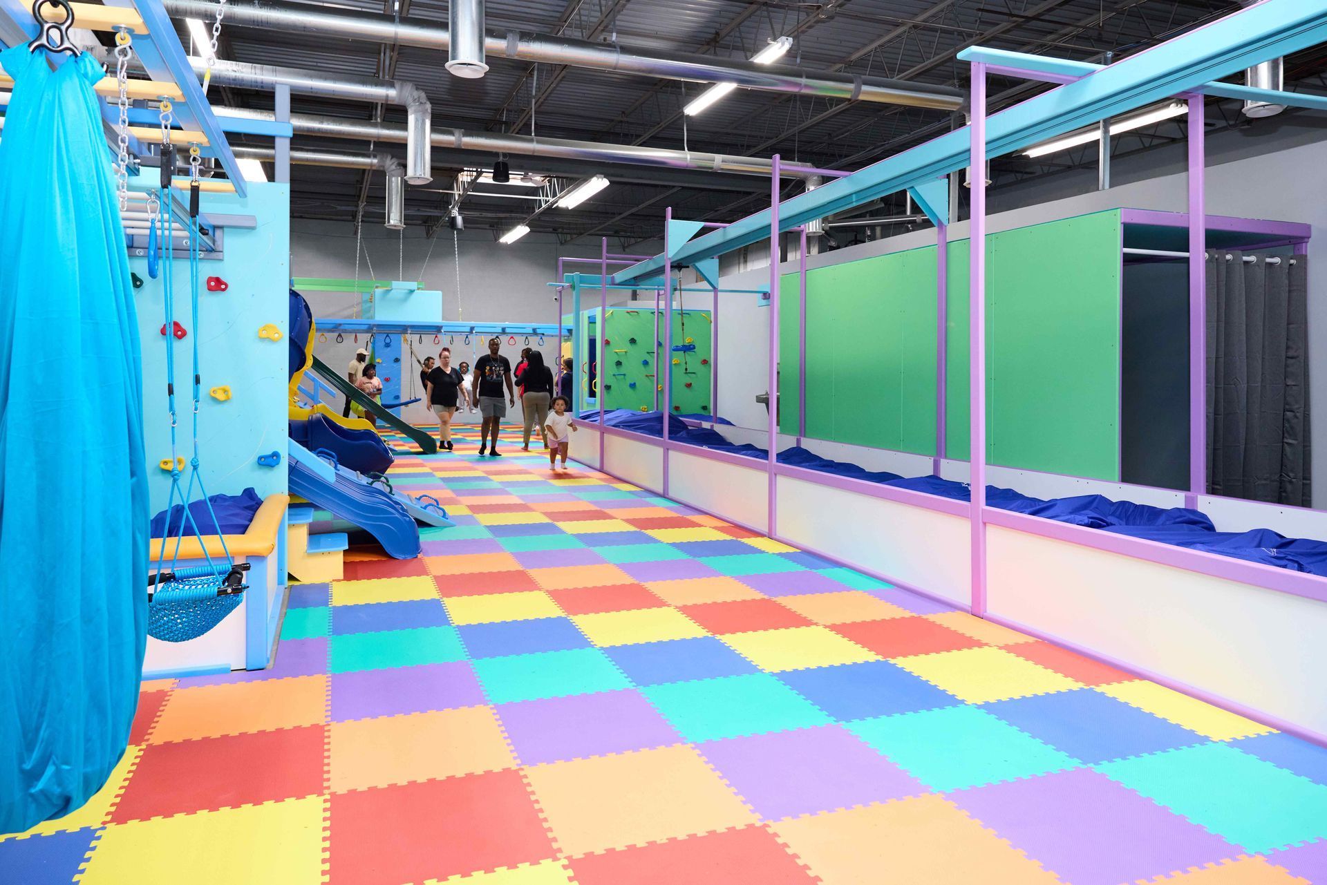 Colorful indoor play area with rainbow floor mats, blue climbing structures, and a few people in the background