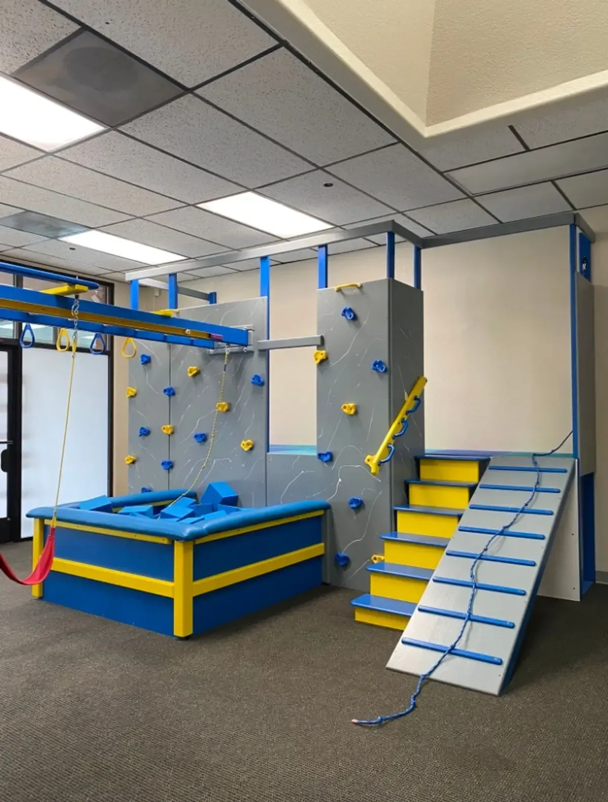 Indoor children's obstacle course with climbing wall, blue padded platforms, and yellow stairs in a playroom