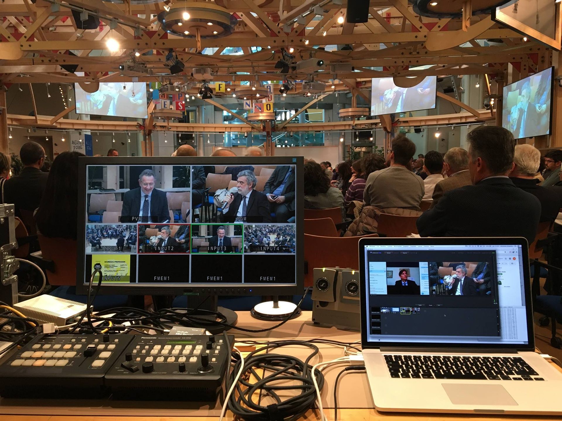 Un evento in streaming live in corso. I monitor mostrano altoparlanti, un mixer audio e un computer portatile che mostra una presentazione in una grande sala.