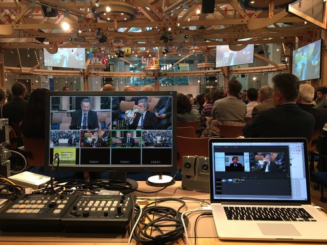 Un evento in streaming live in corso. I monitor mostrano altoparlanti, un mixer audio e un computer portatile che mostra una presentazione in una grande sala.