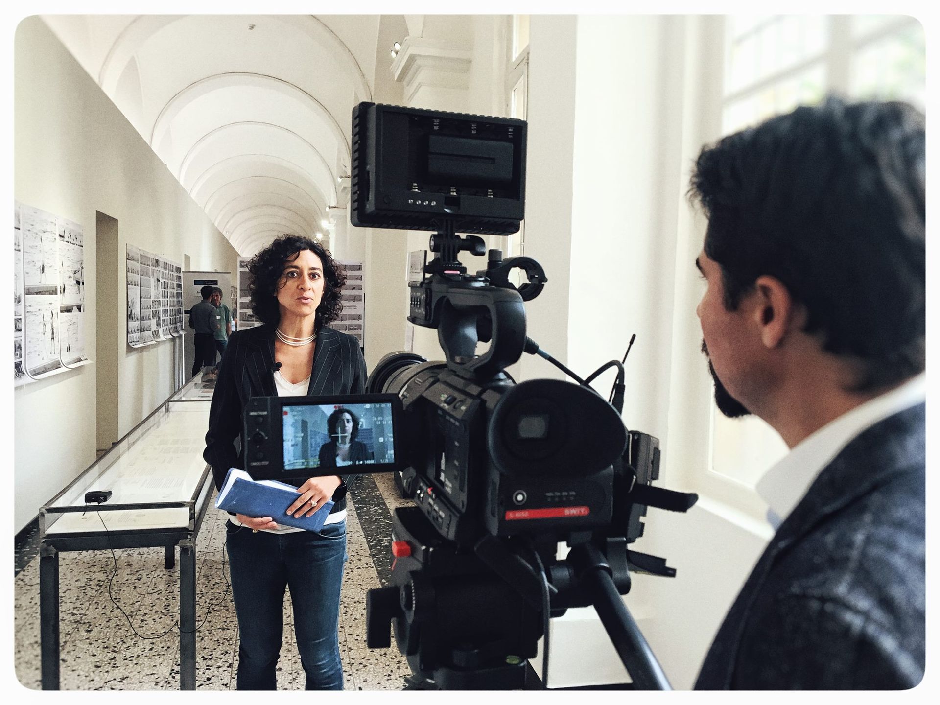 Una donna viene intervistata in un lungo corridoio da una persona con una telecamera su un treppiede.