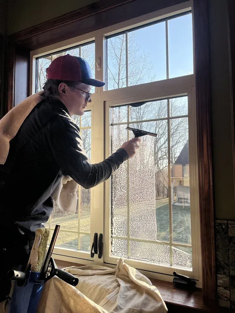 A person cleaning a window with a squeegee. They wear a hat and jacket. The window has a view of a yard and trees.