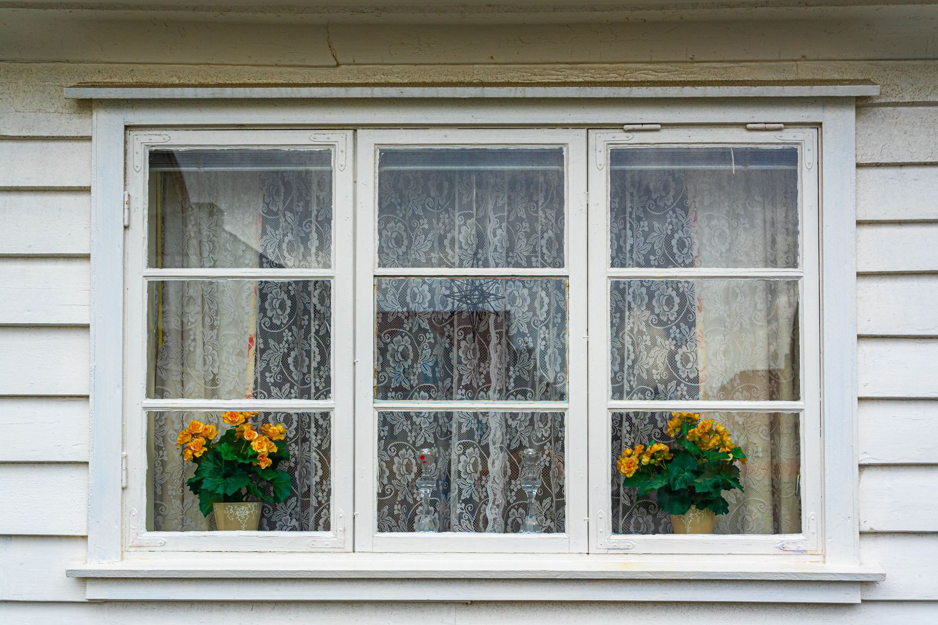 White window with three panes, each containing lace curtains and a pot of yellow flowers.