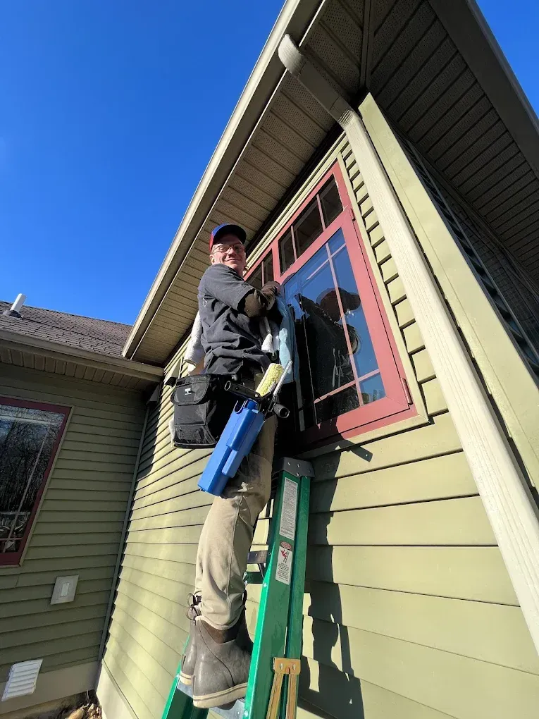 Man cleaning a window on a house exterior using a ladder, with a blue sky background. 