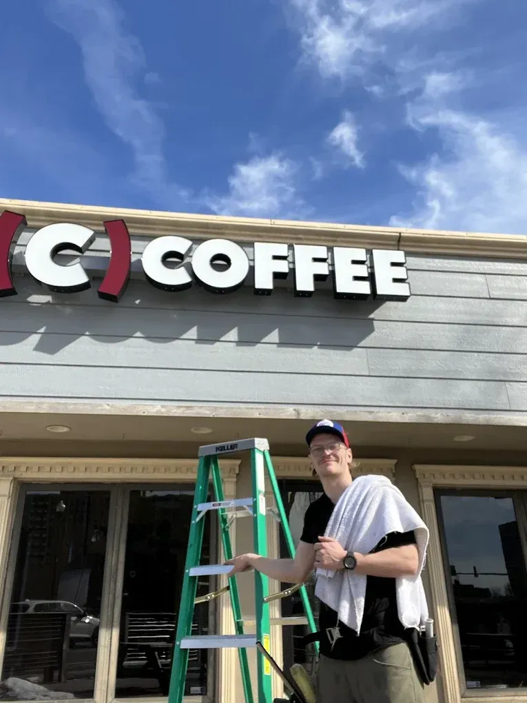 Man on ladder by a store with a "C Coffee" sign, holding a towel, under a blue sky.