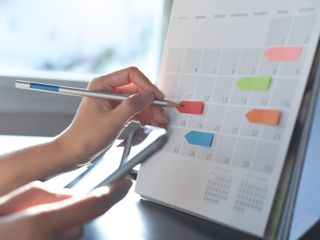 Hand placing a red sticky note on a desk calendar beside a smartphone and pen