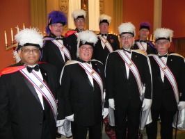 Group of uniformed people in black suits and white plumed hats posing indoors at an event