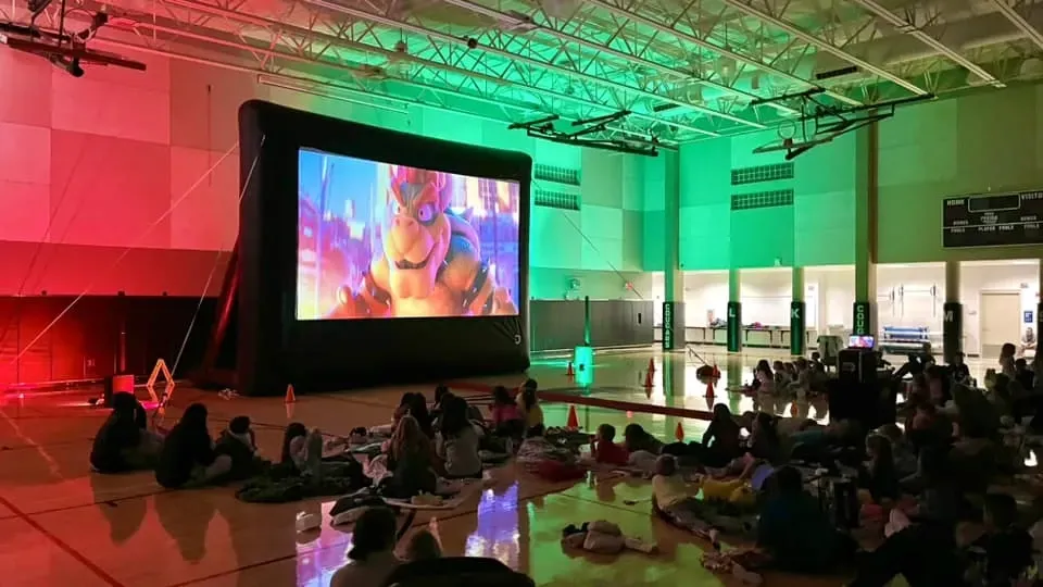 A group of people are watching a movie on a large screen in a gym.
