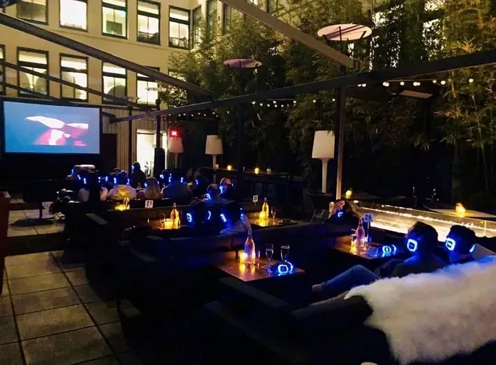 A group of people are sitting at tables in front of an inflatable movie screen watching a movie with silent cinema headphones