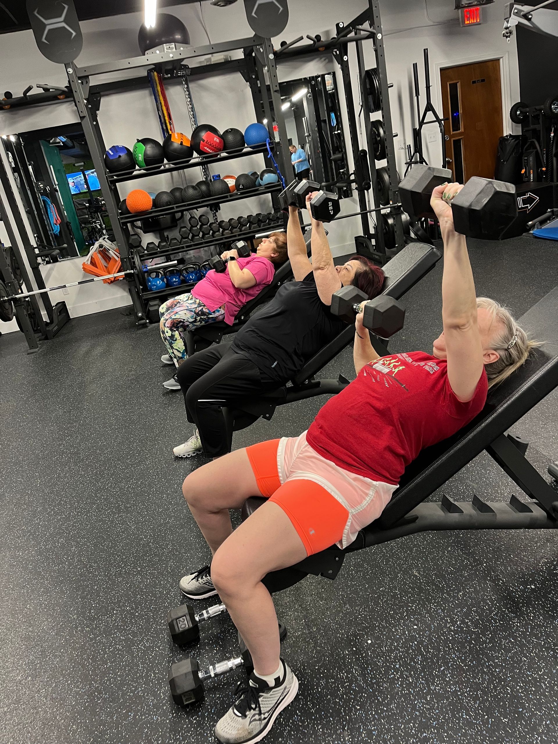 A group of women are sitting on benches in a gym lifting dumbbells.