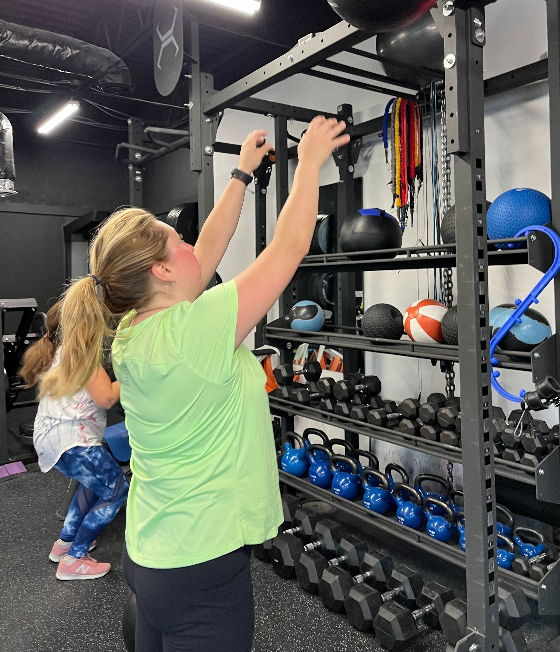 A woman in a green shirt is reaching for a dumbbell in a gym.