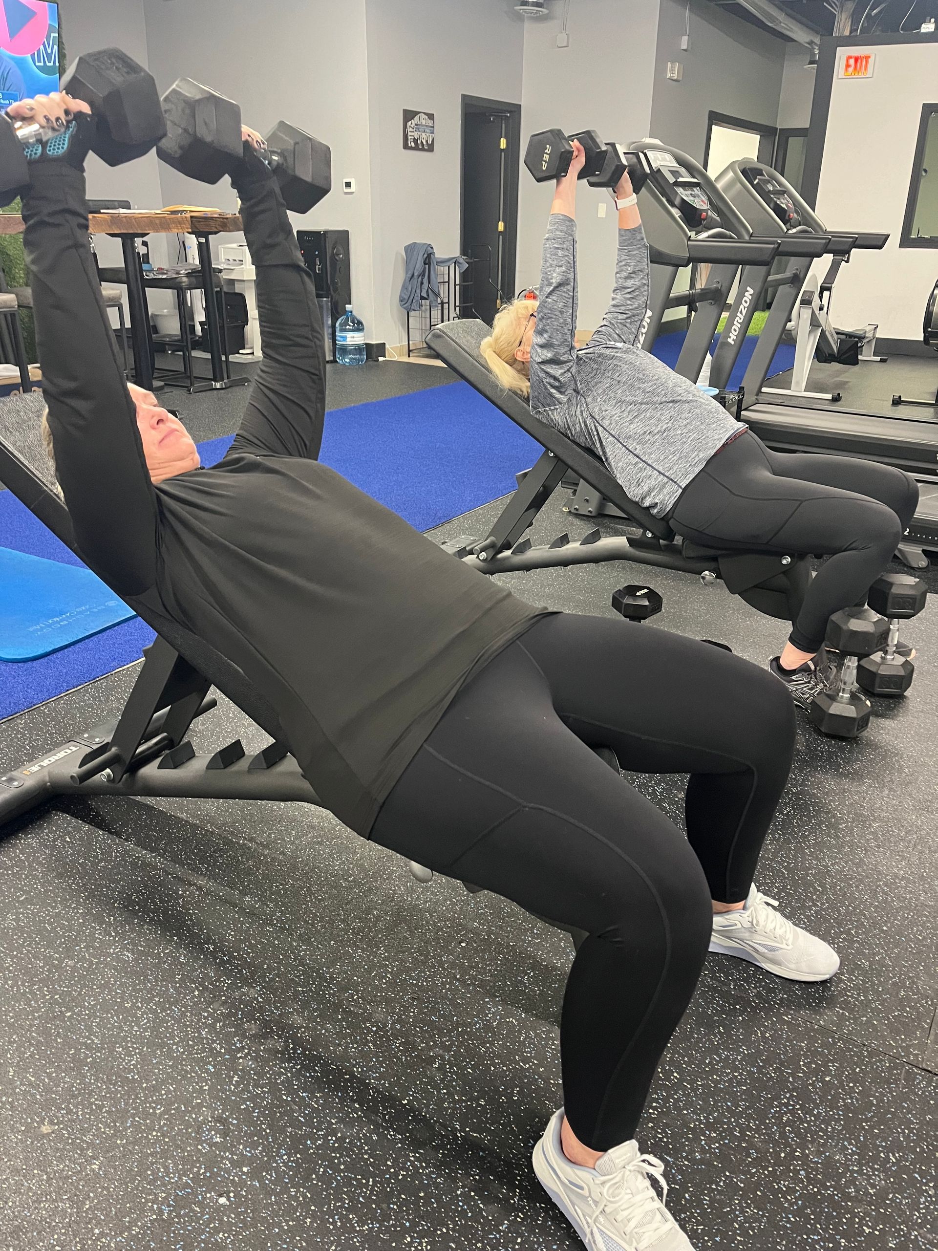 Two women are lifting dumbbells on a bench in a gym.