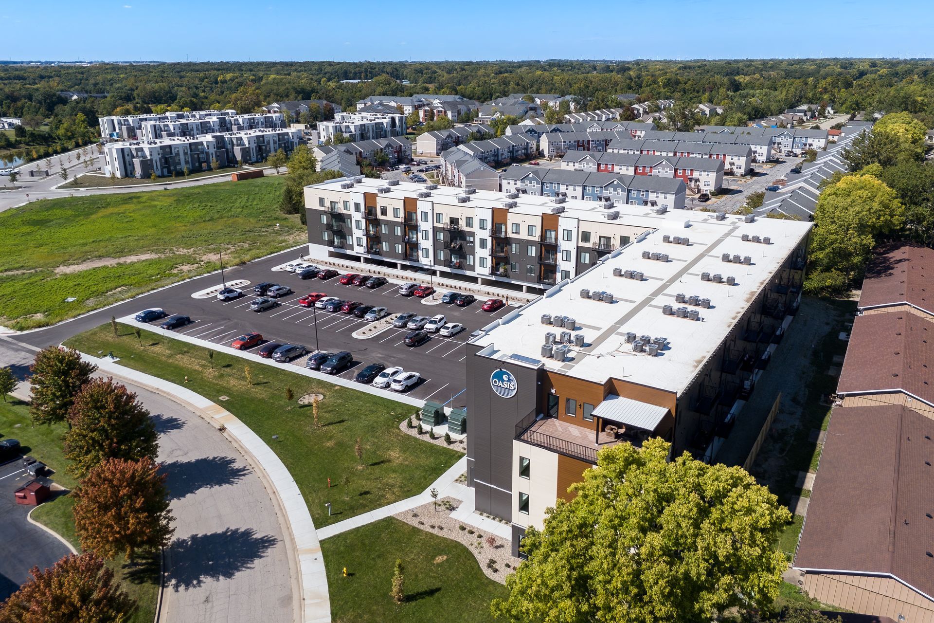 Aerial view of a multi-story apartment complex with parking, trees, and other buildings on a sunny day.