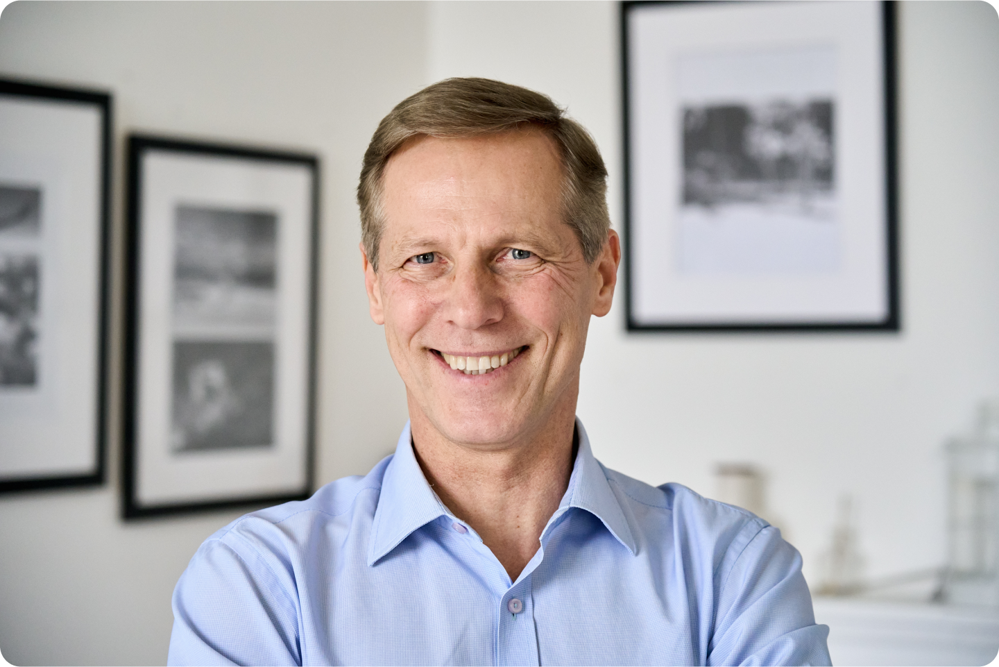 A man in a blue shirt is smiling in front of a wall with pictures on it.