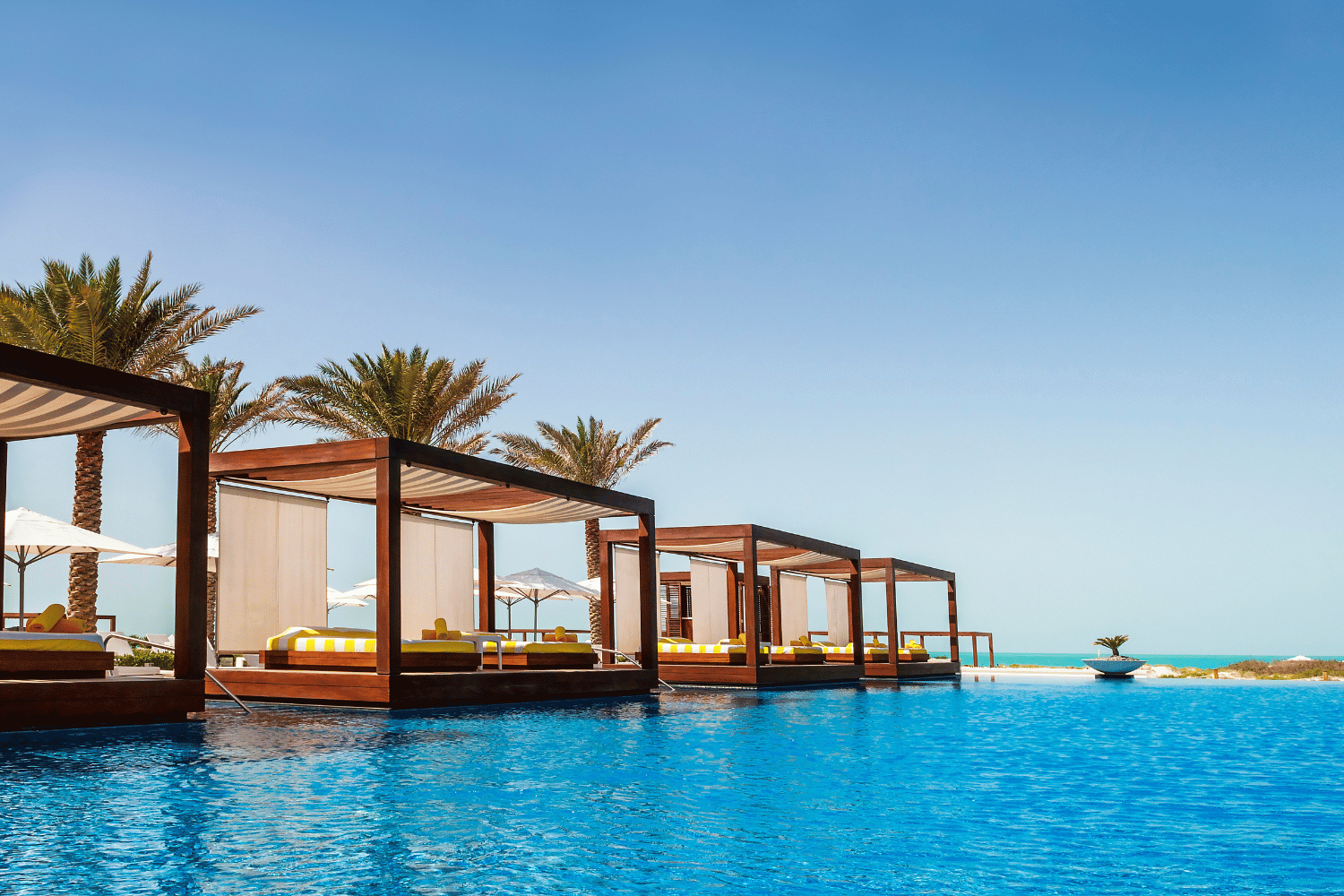Pool with wooden cabanas, palm trees, and ocean under a blue sky.
