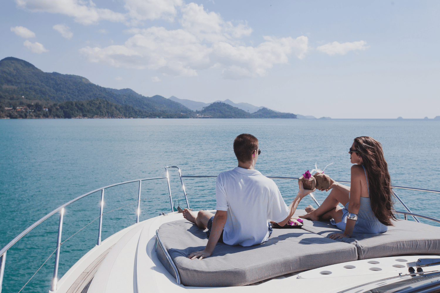 Two people relaxing on a boat deck, facing a calm blue sea with mountains in the distance.