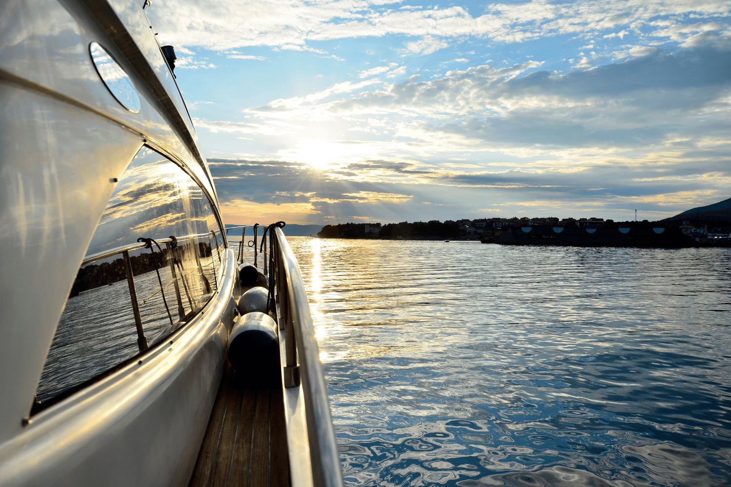 Sailboat deck at sunset with calm water, distant shoreline, and cloudy sky shimmering with golden light
