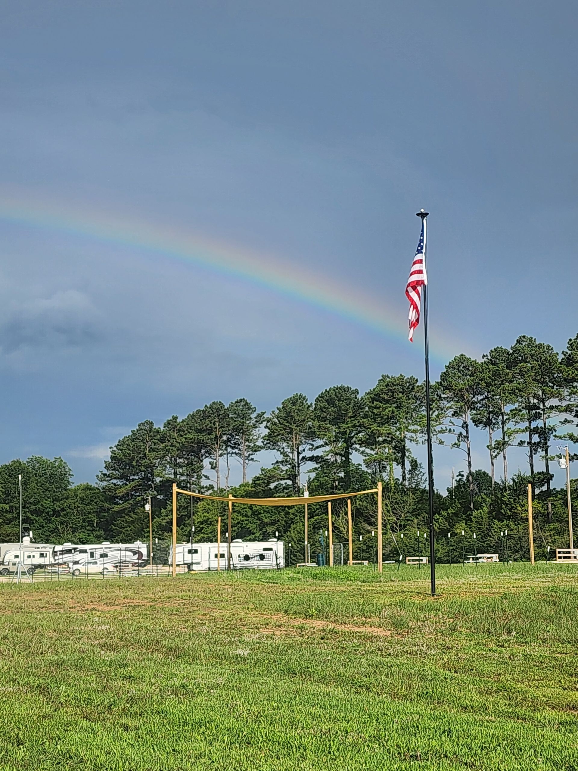 American flag on a pole over a grassy field with trees, white RVs, and a faint rainbow in the sky