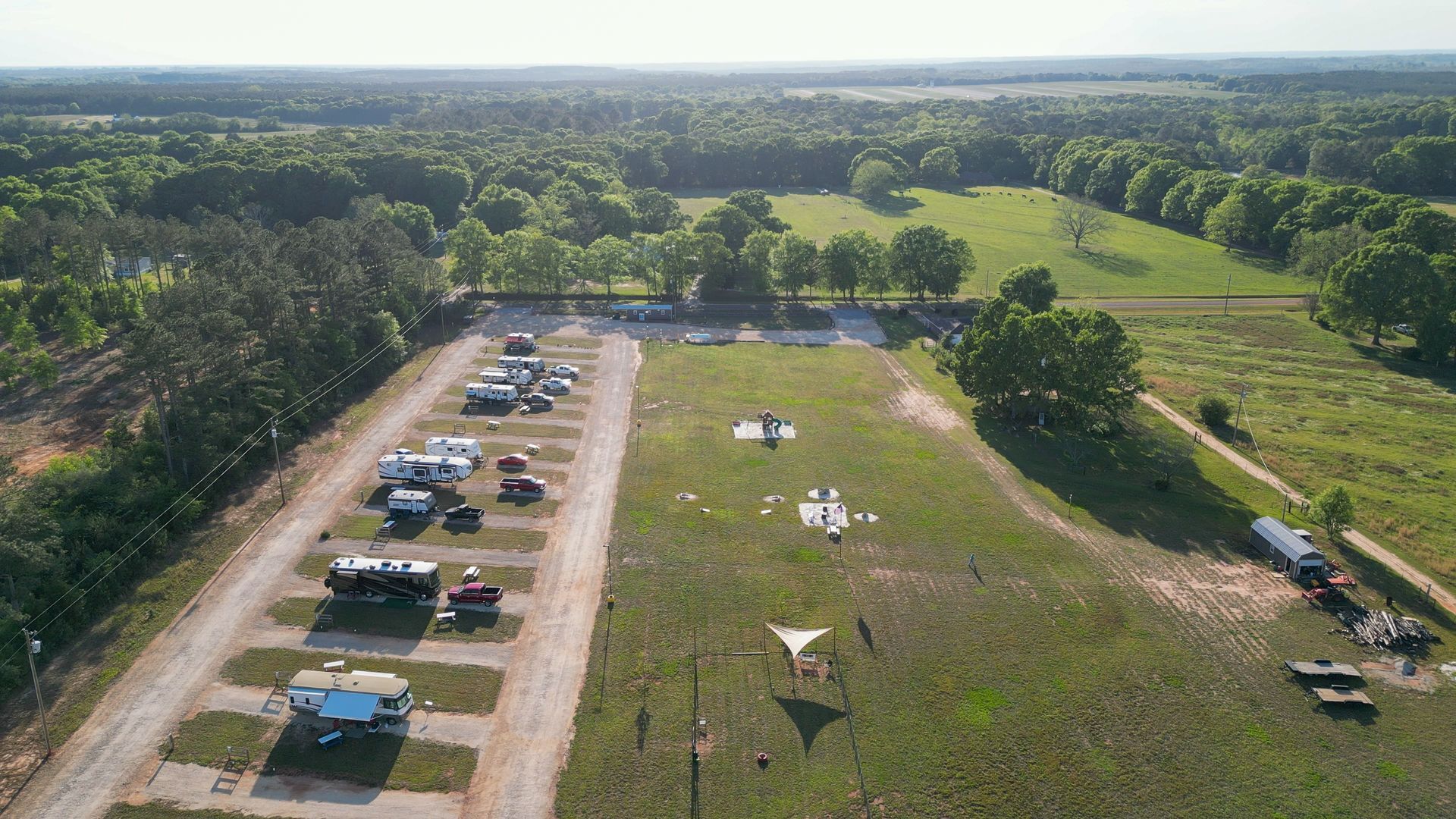 Aerial view of a grassy field beside parked cars and tents, with trees and open countryside beyond.