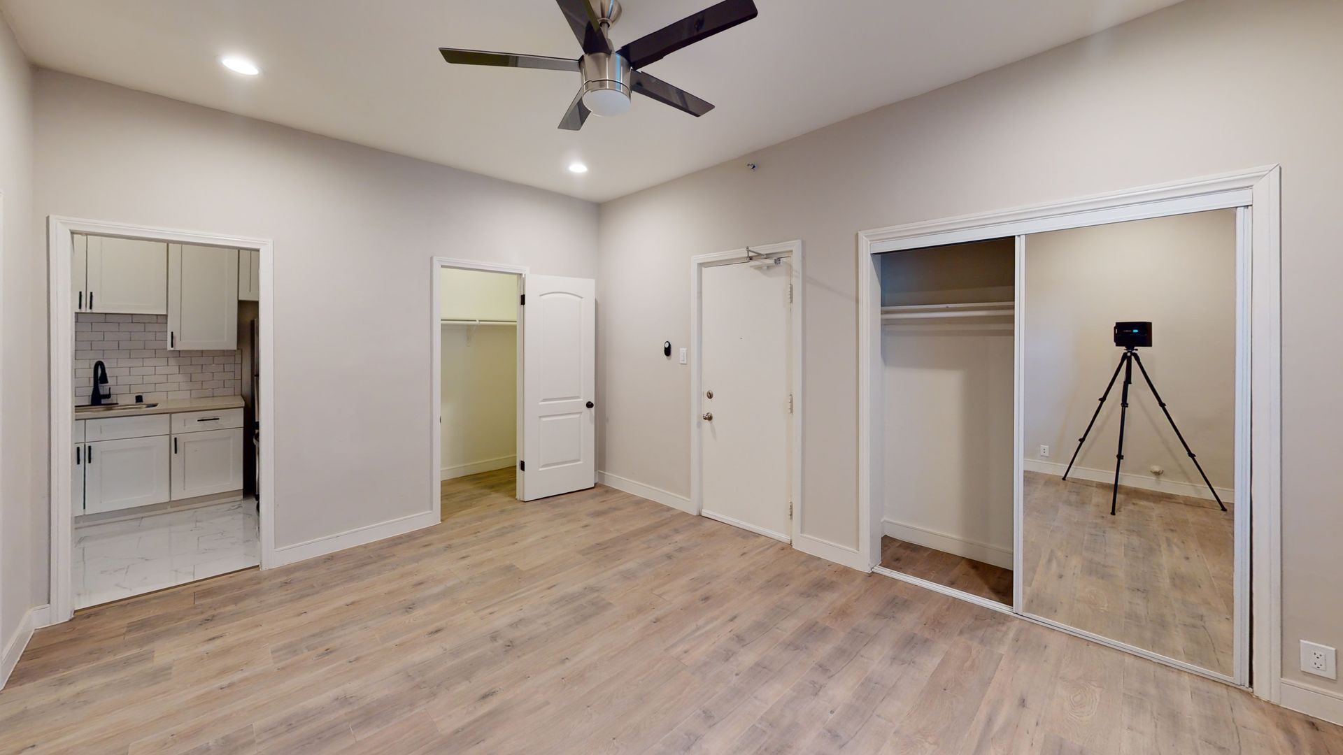 Empty room with light wood floors, a closet, a doorway to a kitchen, and a camera on a tripod.